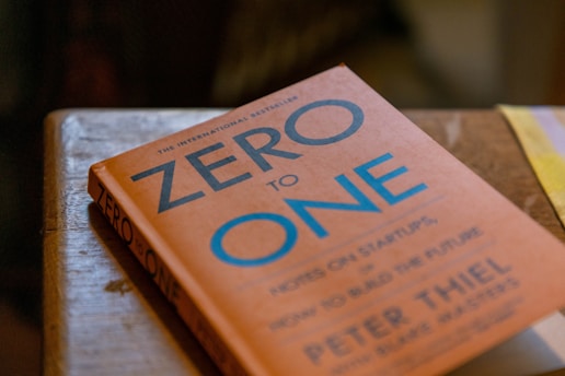 A book titled 'Zero to One' is resting on a wooden surface. The cover is orange with blue and white text, and the author is Peter Thiel. There is soft lighting, and the book's edges are slightly worn, suggesting it has been read or handled.