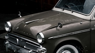 An antique car with a classic design is parked, featuring a glossy dark olive paint job. The vehicle has dual round headlights and a chrome grille. The vintage aesthetics are highlighted by the metallic trim and round side mirrors.