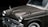 An antique car with a classic design is parked, featuring a glossy dark olive paint job. The vehicle has dual round headlights and a chrome grille. The vintage aesthetics are highlighted by the metallic trim and round side mirrors.