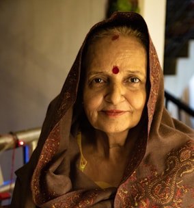 An elderly woman wearing a traditional brown shawl with intricate patterns sits indoors. A red bindi is prominently placed on her forehead, and she gazes warmly at the camera. Her surroundings include a staircase and soft lighting that creates a cozy atmosphere.