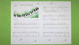 A sheet of classical music titled 'Classical Hodgepodge' is displayed, featuring a whimsical illustration of a piano keyboard flowing through a grassy landscape. The sheet contains musical notes and compositions on a white background, with additional decorative musical notes around the edges. The pages are set against a light green textured background.