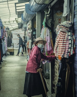 An alleyway lined with clothing stalls features an elderly woman wearing a mask and hat, holding a cane, examining items for sale. Another person, also masked, walks further down the aisle carrying a shopping bag. The atmosphere is dimly lit with assorted garments hanging prominently on both sides.