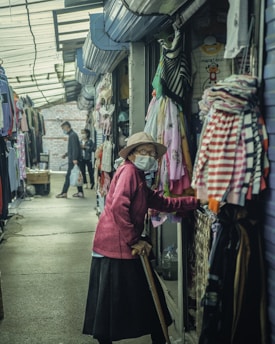 An alleyway lined with clothing stalls features an elderly woman wearing a mask and hat, holding a cane, examining items for sale. Another person, also masked, walks further down the aisle carrying a shopping bag. The atmosphere is dimly lit with assorted garments hanging prominently on both sides.