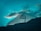 A majestic snow-capped mountain peak against a vibrant teal sky, partially covered by soft clouds.