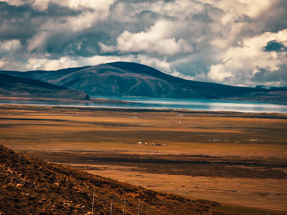 brown field near mountain under white clouds and blue sky during daytime