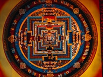 Intricate mandala art featuring concentric geometric patterns in vivid colors. Symbols and ornate scripts decorate the circular form, with dominant blue, orange, and red tones enhancing its complexity and depth.