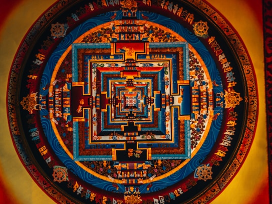 Intricate mandala art featuring concentric geometric patterns in vivid colors. Symbols and ornate scripts decorate the circular form, with dominant blue, orange, and red tones enhancing its complexity and depth.