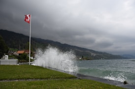A large, crashing wave hits the shore near a grassy area with a Swiss flag waving on a flagpole. The sky is overcast and dark, suggesting an impending storm. In the background, there are hilly areas covered with trees and a cluster of buildings.