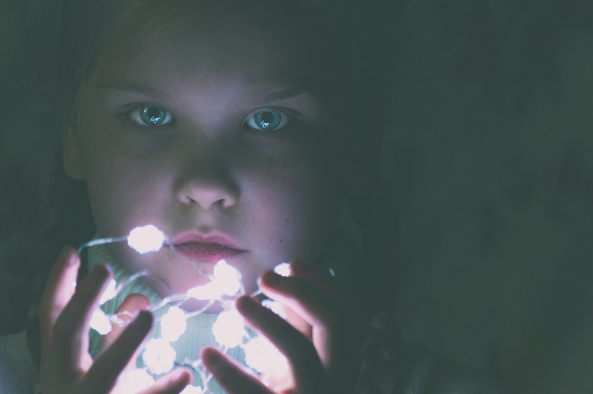 A young child is holding a string of glowing fairy lights close to their face, casting a soft, ethereal glow. The light illuminates their facial features, creating an atmosphere of curiosity and wonder.