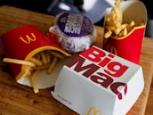 A wooden surface is topped with fast food items including two packages of French fries in red packaging, a wrapped bacon burger in purple wrapping, and a closed Big Mac box. The scene is typical of a fast-food meal setting.