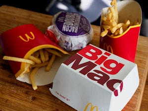 A wooden surface is topped with fast food items including two packages of French fries in red packaging, a wrapped bacon burger in purple wrapping, and a closed Big Mac box. The scene is typical of a fast-food meal setting.