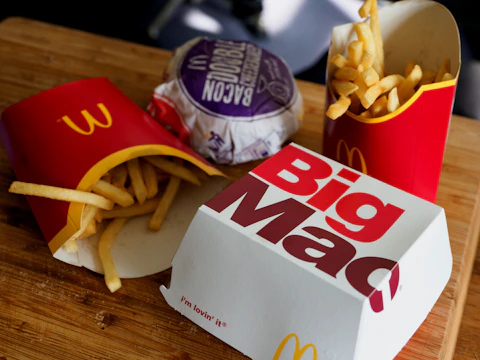 A wooden surface is topped with fast food items including two packages of French fries in red packaging, a wrapped bacon burger in purple wrapping, and a closed Big Mac box. The scene is typical of a fast-food meal setting.