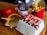 A wooden surface is topped with fast food items including two packages of French fries in red packaging, a wrapped bacon burger in purple wrapping, and a closed Big Mac box. The scene is typical of a fast-food meal setting.