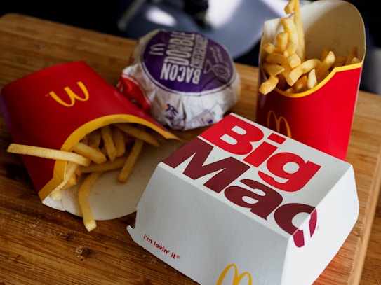 A wooden surface is topped with fast food items including two packages of French fries in red packaging, a wrapped bacon burger in purple wrapping, and a closed Big Mac box. The scene is typical of a fast-food meal setting.