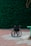 A lone black wheelchair is situated on a tiled area with red and grey square tiles. In the background, there is a lush green hedge that covers the entire width of the image. There are some scattered dry leaves on the ground and a partial view of a green bench on the right-hand side.
