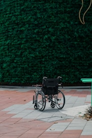 A lone black wheelchair is situated on a tiled area with red and grey square tiles. In the background, there is a lush green hedge that covers the entire width of the image. There are some scattered dry leaves on the ground and a partial view of a green bench on the right-hand side.
