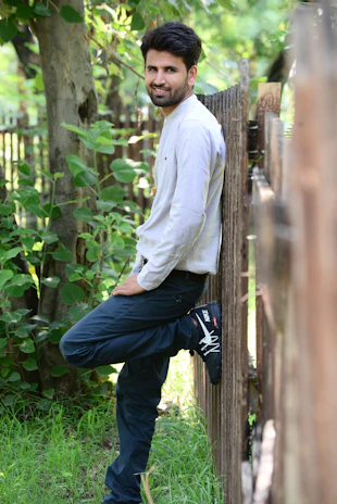 Casual daytime photo of the model smiling while leaning on a wooden fence outdoors.