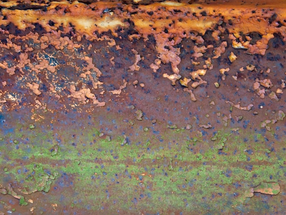 A close-up view of a metal surface with extensive rust and corrosion. The texture is rough, with flaking and peeling layers of rust in shades of orange, brown, and green. Patches of darker spots are scattered throughout.