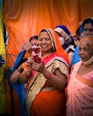 A group of women, dressed in colorful traditional Indian sarees, are gathered together. One woman in the foreground, wearing an orange saree with gold embroidery, is smiling as she looks at a smartphone adorned with a floral case. The women behind her are wearing sarees in various colors, including blue and pink, and some have face masks on. The background features bright orange and yellow drapes.