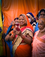 A group of women, dressed in colorful traditional Indian sarees, are gathered together. One woman in the foreground, wearing an orange saree with gold embroidery, is smiling as she looks at a smartphone adorned with a floral case. The women behind her are wearing sarees in various colors, including blue and pink, and some have face masks on. The background features bright orange and yellow drapes.