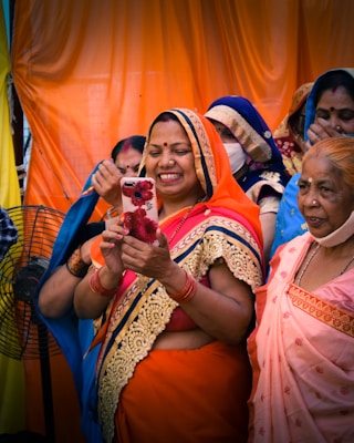 A group of women, dressed in colorful traditional Indian sarees, are gathered together. One woman in the foreground, wearing an orange saree with gold embroidery, is smiling as she looks at a smartphone adorned with a floral case. The women behind her are wearing sarees in various colors, including blue and pink, and some have face masks on. The background features bright orange and yellow drapes.