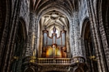 A grand, ornate pipe organ is situated in the interior of a historic cathedral, featuring high vaulted ceilings made of stone arches. The organ is flanked by statues, and natural light streams in through a stained glass window above, casting shadows across the weathered stone walls.