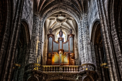 A grand, ornate pipe organ is situated in the interior of a historic cathedral, featuring high vaulted ceilings made of stone arches. The organ is flanked by statues, and natural light streams in through a stained glass window above, casting shadows across the weathered stone walls.