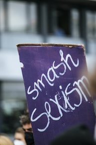 A protest sign with the words 'smash sexism' written in white on a purple background. The sign is held amidst a blurred crowd, suggesting a demonstration or rally.