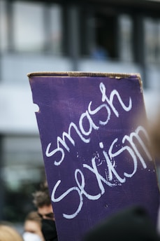 A protest sign with the words 'smash sexism' written in white on a purple background. The sign is held amidst a blurred crowd, suggesting a demonstration or rally.