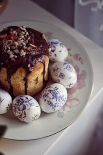 A decorated Easter cake topped with chocolate glaze and chopped nuts is surrounded by intricately painted white Easter eggs. The eggs feature detailed blue floral and bird designs. Everything is presented on a floral-patterned porcelain plate, which rests on a light-colored surface.
