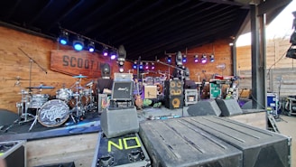 A well-equipped music stage featuring various musical instruments and equipment. On the left, a drum kit with 'Kash'd Out' written on the bass drum is visible. In the center, there are multiple amplifiers, speakers, and microphones. Stage lighting hangs above, illuminating the area with blue and purple hues. The wooden backdrop includes a sign for 'Scootin' since 1871'.