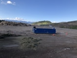 A spacious outdoor area with a large blue dumpster positioned on the ground. To the left, there is a pile of mixed debris or wood, indicative of a construction or disposal site. The background features rolling hills with patches of green grass and a slightly cloudy blue sky. There are cones on the ground indicating caution in the area.