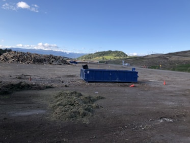 A spacious outdoor area with a large blue dumpster positioned on the ground. To the left, there is a pile of mixed debris or wood, indicative of a construction or disposal site. The background features rolling hills with patches of green grass and a slightly cloudy blue sky. There are cones on the ground indicating caution in the area.