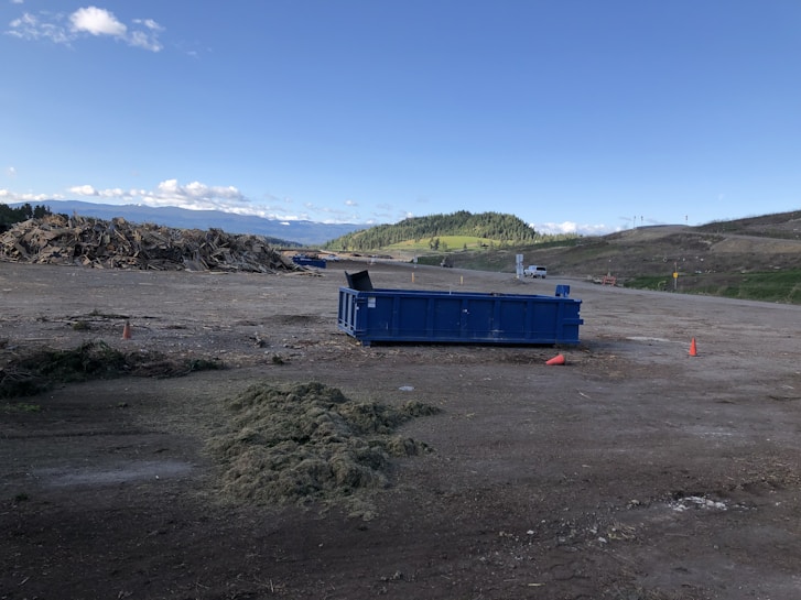 A spacious outdoor area with a large blue dumpster positioned on the ground. To the left, there is a pile of mixed debris or wood, indicative of a construction or disposal site. The background features rolling hills with patches of green grass and a slightly cloudy blue sky. There are cones on the ground indicating caution in the area.