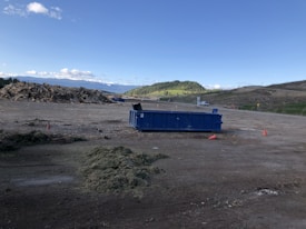 A spacious outdoor area with a large blue dumpster positioned on the ground. To the left, there is a pile of mixed debris or wood, indicative of a construction or disposal site. The background features rolling hills with patches of green grass and a slightly cloudy blue sky. There are cones on the ground indicating caution in the area.