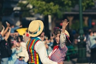 A dancing couple dressed in brightly colored vintage clothing in an outdoor setting. The woman is wearing a blue and red patterned dress with a matching hat, while the man has a striped vest and a straw boater hat. They are smiling and engaging with a lively crowd in the background.