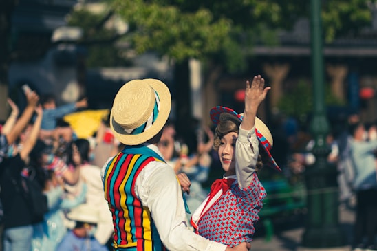 A dancing couple dressed in brightly colored vintage clothing in an outdoor setting. The woman is wearing a blue and red patterned dress with a matching hat, while the man has a striped vest and a straw boater hat. They are smiling and engaging with a lively crowd in the background.
