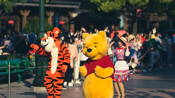 A colorful scene featuring two people dressed in character costumes, one as a yellow bear with a red shirt and the other as an orange and black tiger. They are in an amusement park setting, surrounded by a crowd of visitors. The environment is lively and festive, with people in the background taking photos and enjoying themselves.