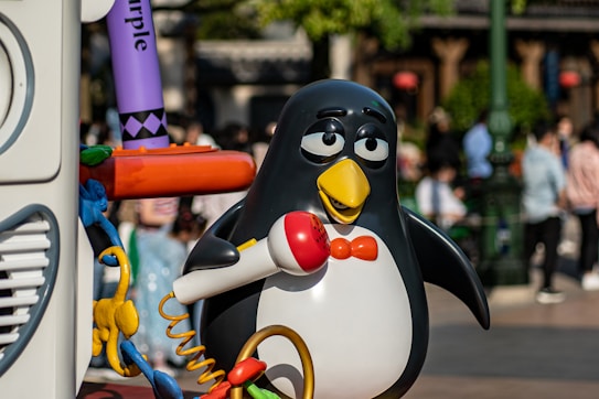 A cartoonish penguin character with a yellow beak, red bow tie, and red microphone stands next to a colorful setup. The scene is outdoors with several people in the background, slightly out of focus.