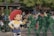 A person wearing a costume of a cartoon character with a large red hat and detailed outfit stands in a public setting. Nearby, three performers are dressed as toy soldiers in solid green uniforms. They are part of a lively outdoor atmosphere with trees and various people in the background, some holding umbrellas.