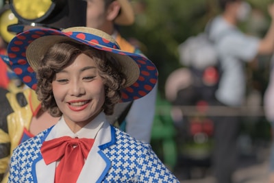 A person wearing a colorful, vintage-style outfit with a blue and white patterned jacket and a large brimmed hat adorned with blue and red polka dots. The person has curly brown hair and is smiling warmly. In the background, there are blurred people and a person in a costume with yellow accents.