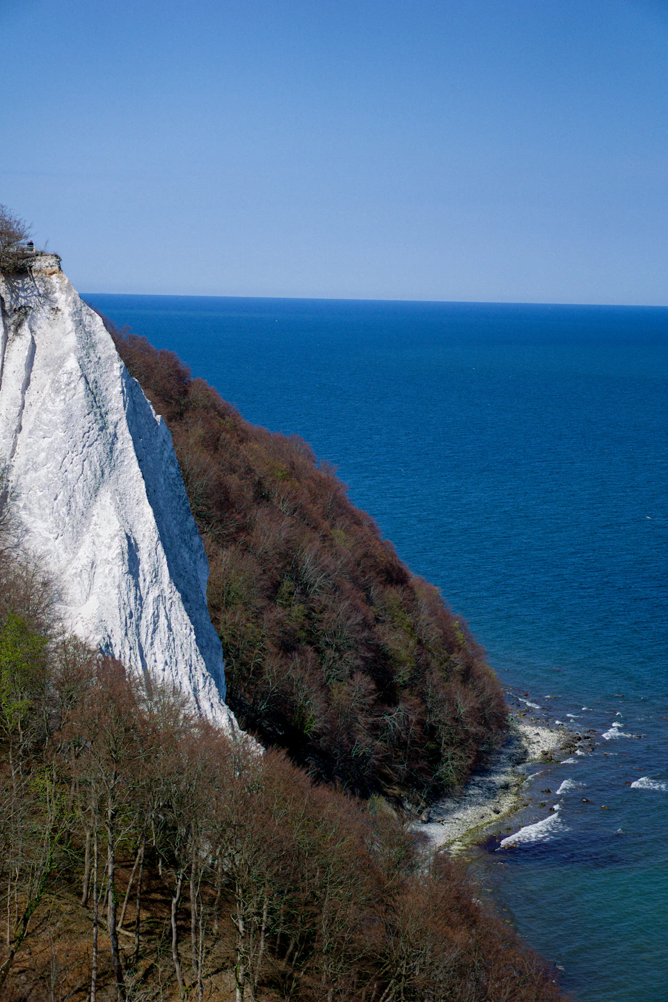 The image depicts a cliff face with white chalk lines, showcasing the rugged beauty of nature. The cliff is situated on the edge of a body of water, creating a striking contrast between the rocky terrain and the blue expanse below. The perspective of the photo is from above, providing an expansive view of the cliff face and its surroundings. In the distance, trees can be seen peeking out from behind the cliff, adding depth to the scene.