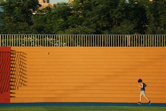 A person in athletic gear is jogging in front of a large orange wall with horizontal steps. The wall casts long shadows, and there are trees visible above the wall, creating a natural background.