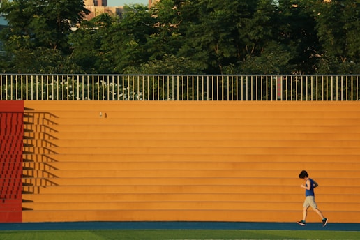 A person in athletic gear is jogging in front of a large orange wall with horizontal steps. The wall casts long shadows, and there are trees visible above the wall, creating a natural background.