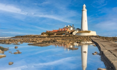A tall white lighthouse stands on a rocky shoreline surrounded by water with reflections visible on the surface. Nearby, there are several small buildings with red roofs, likely cottages or service buildings, sitting on the rugged terrain. The sky is clear with a blue hue, indicating a bright, sunny day.