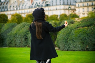 A person wearing a black coat and hood is feeding birds in a garden. The background shows a historic building with trees and neatly trimmed hedges alongside a well-maintained lawn.