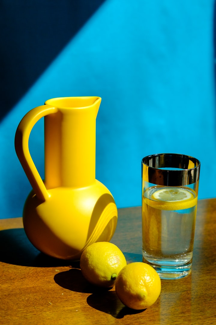 A yellow jug placed to the left, with a rounded bottom and a cylindrical top. Two round lemons placed one before the other t
