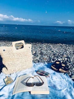 A serene beach setting with a woven bag and a pair of sandals on a pebbled shore. An open book with sunglasses resting on it lies on a light blue cloth. The background features a calm sea and clear blue sky with a few clouds.