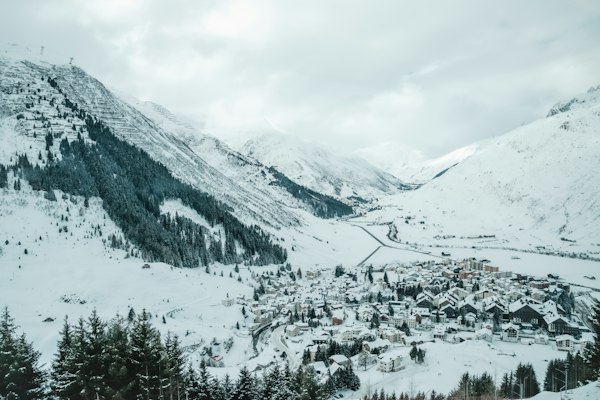 Snow-covered Andermatt village and the flat white floor of the Urseren valley seen from above, with snowy conifers in the foreground
