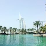 A large sail-shaped building, surrounded by palm trees, stands beside a tranquil body of water on a bright, sunny day. The structure is well-known for its modern and unique architectural design.
